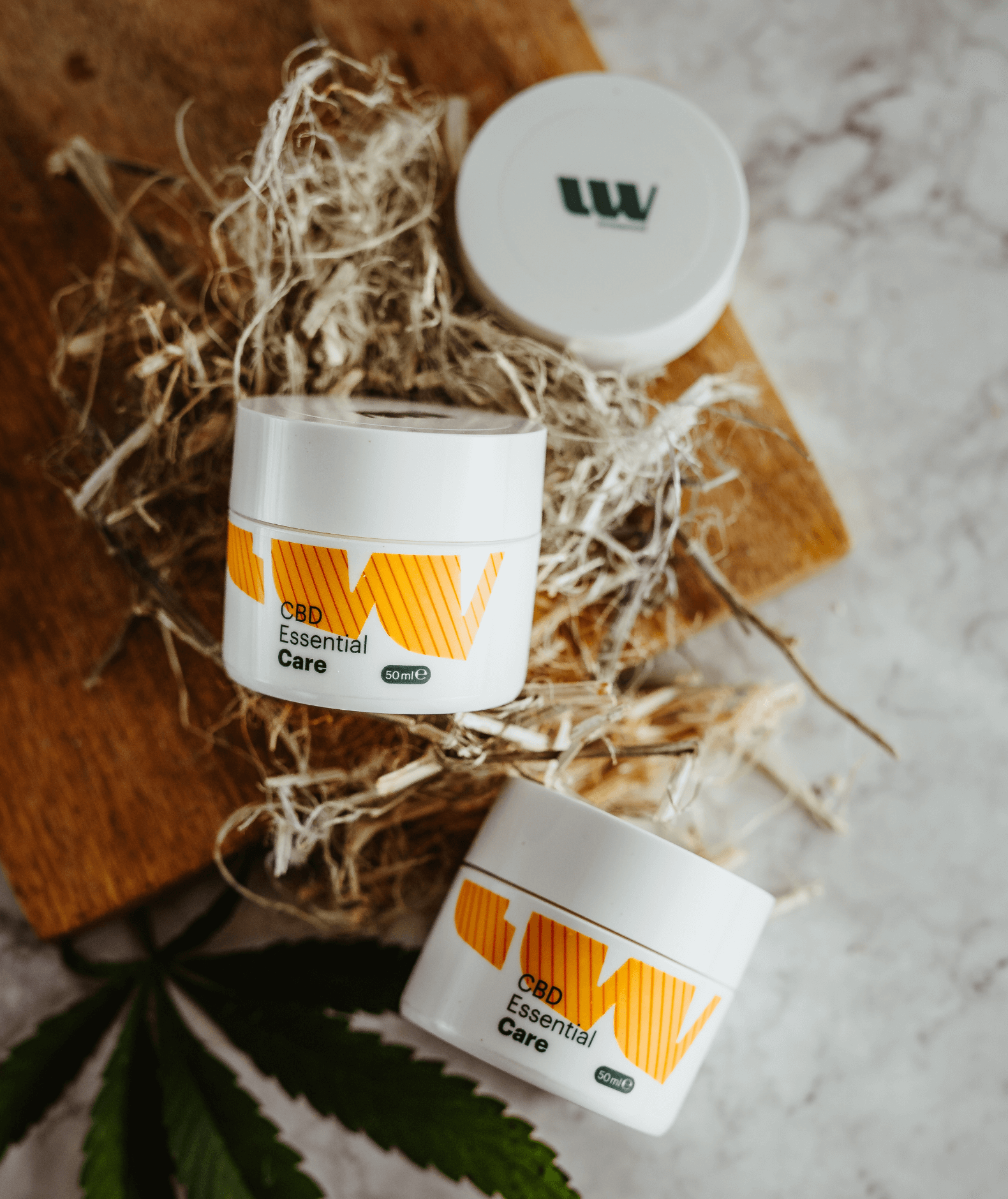 Zwei Creme-Tiegel der CBD Essential Care auf Holzbrett mit Hanfblatt